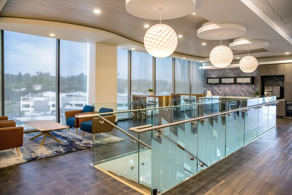 A modern office lounge with large windows, stylish furniture, round hanging lights from a recent interior lighting installation, glass railings, and a mix of blue and brown chairs around wooden tables on a dark wood floor.